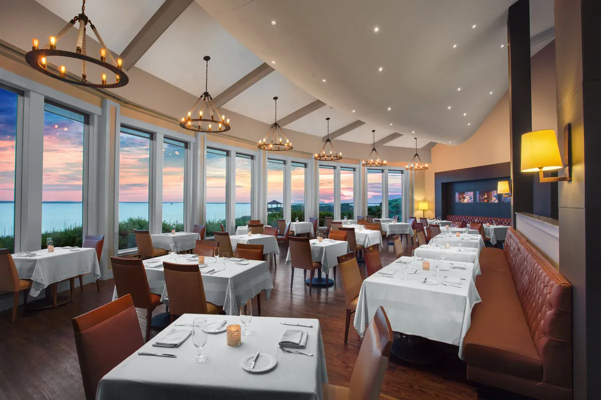 An elegant interior dining space is showcased with large windows framing a stunning sunset view. Tastefully set tables and soft chandeliers create an intimate, sophisticated atmosphere suited for romantic fine dining experiences.