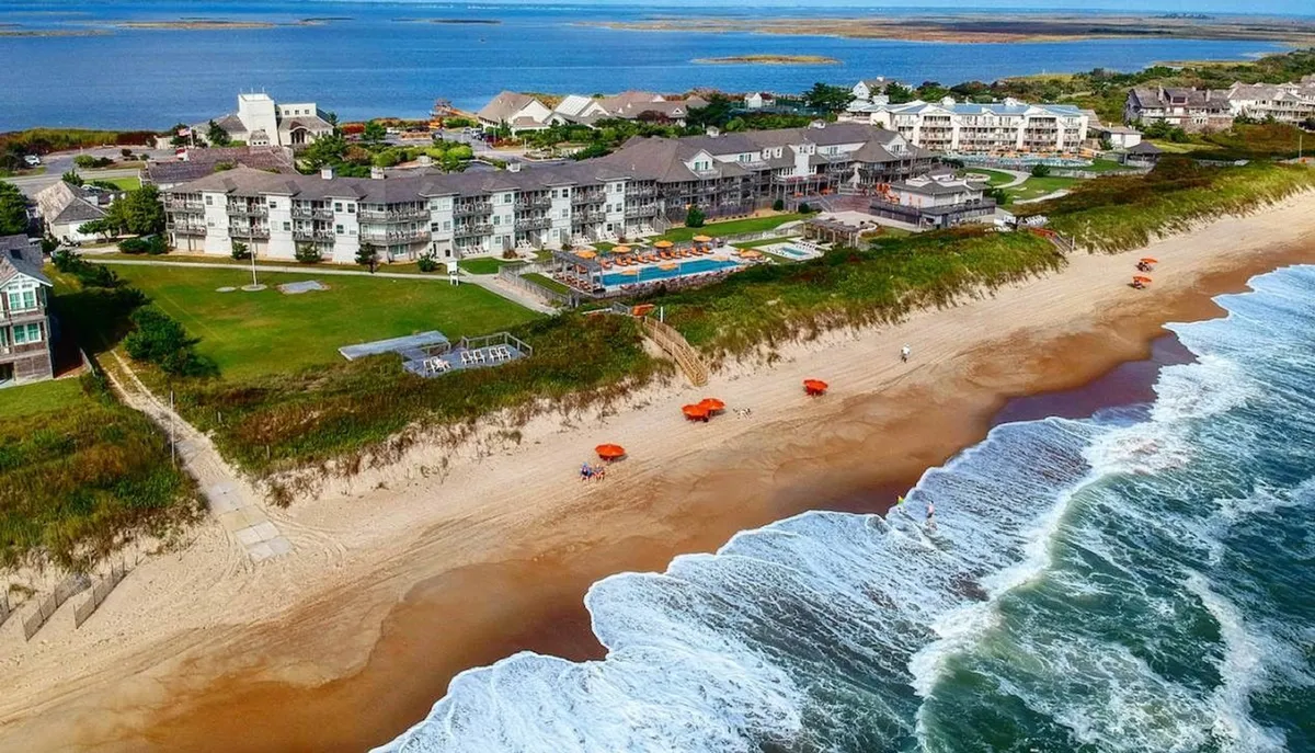 A vibrant exterior shot showcasing the resort as a true beachfront property, with a sparkling swimming pool in the foreground, umbrellas dotting both poolside and beach areas, and gentle ocean waves beyond. Families can be seen enjoying both the sand and the pool, creating a lively, welcoming atmosphere.