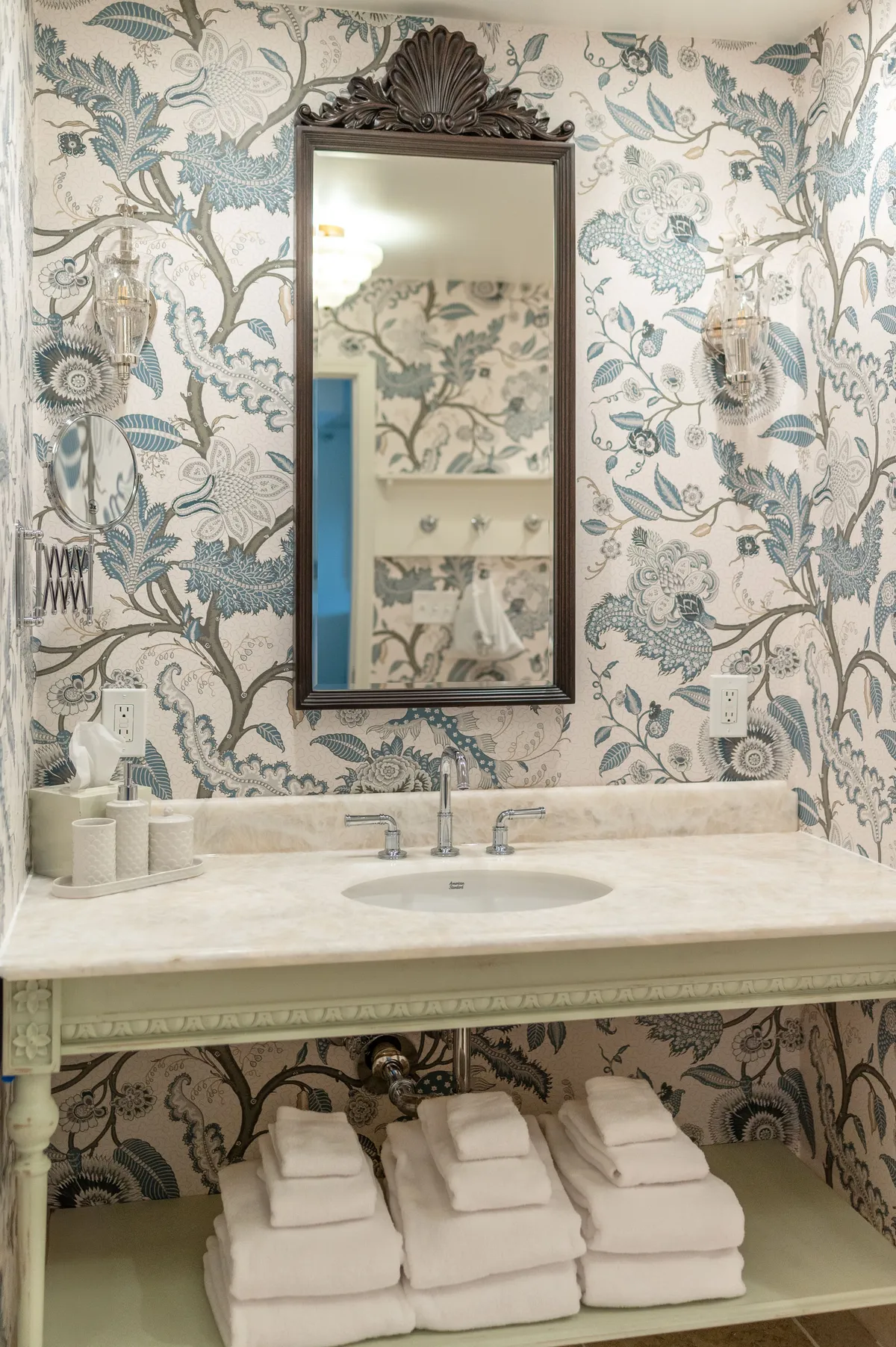 A serene and elegant bathroom scene featuring an ornate mirror above a sink with a marble countertop. Neatly stacked towels and soft, floral wallpaper add to the soothing ambiance. The setting is inviting for personal grooming and relaxation, with a subtle hint of luxury and attention to detail in the fixtures and décor.