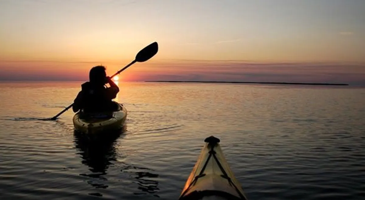 A silhouette of a kayaker glides across calm water with the sun setting in the background, painting the sky in rich hues. The water reflects the stunning sunset, suggesting peacefulness and a strong connection to the natural beauty that surrounds the resort.