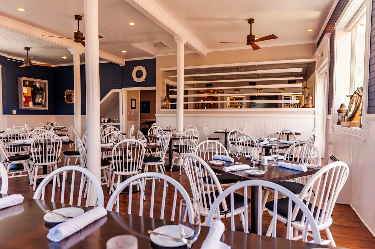 A welcoming and relaxed interior restaurant featuring white wooden chairs, dark wooden tables, decorative wall art, and a cozy bar area. The space is designed for guests to gather, dine, and celebrate special occasions, with an atmosphere of warmth and casual sophistication.
