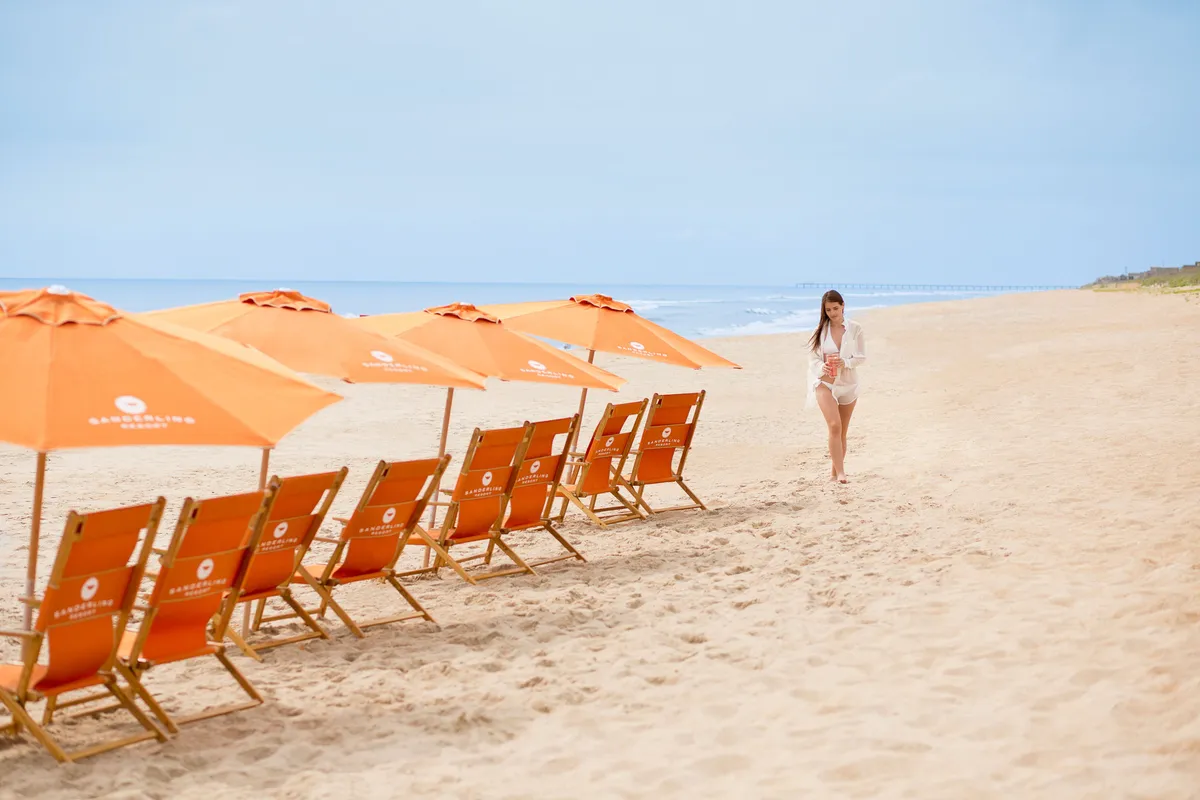 A serene beachfront scene with beach chairs and umbrellas facing the ocean. A woman strolls along the waterline, while the sun is low in the sky, setting a tranquil mood. The arrangement of chairs suggests a perfect spot to unwind and soak up ocean panoramas, with broad views of both sea and sky.