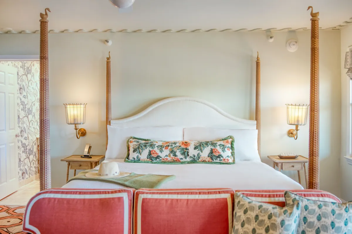 A tranquil studio space with a four-poster bed dressed in luxury linens, complemented by a floral decorative pillow and bedside lamps. The intricate wallpaper and soft lighting evoke a sense of warmth and relaxation, perfect for unwinding after a day in the sun.