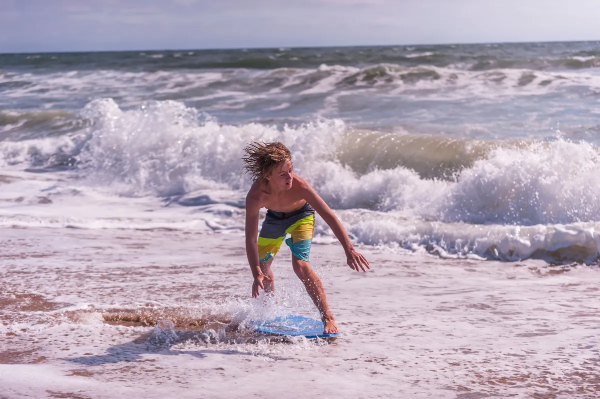 A joyful action shot of a young boy riding a boogie board on the ocean waves. The bright sunlight, splashing water, and visible shoreline convey excitement, play, and family-friendly beach activities.