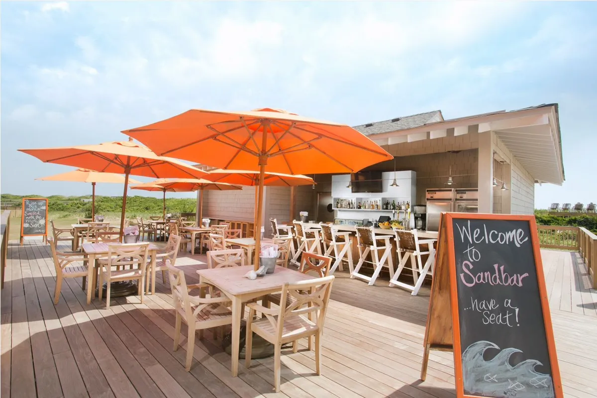 A lively outdoor dining area under orange umbrellas, with wooden tables and a sandbar sign marking the casual bar. Guests gather to enjoy food, drinks, and conversation, while the open-air setup and sunset ambiance capture the relaxed, social spirit of the resort’s dining scene.