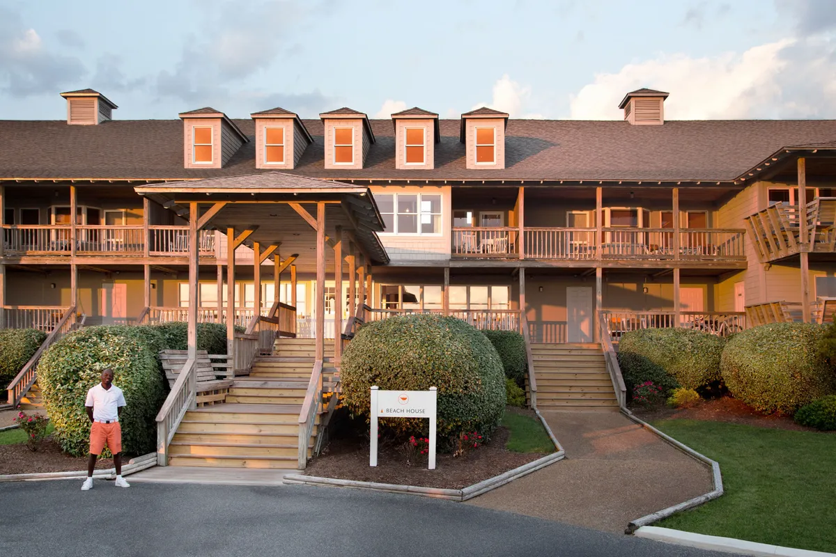 This exterior image captures the inviting entrance of the beach house, landscaped bushes, and a relaxed figure standing out front. The porch area is visible, suggesting spaces for gathering, relaxing, and enjoying outdoor activities, while the overall scene is bathed in the warm, tranquil light of sunset.