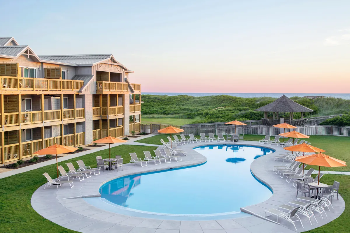 An inviting pool scene with families swimming, sunbathing under umbrellas, and mingling near the landscaped garden. The resort’s building facade provides a backdrop, and the mood is social and relaxed, suggesting group games and gatherings.