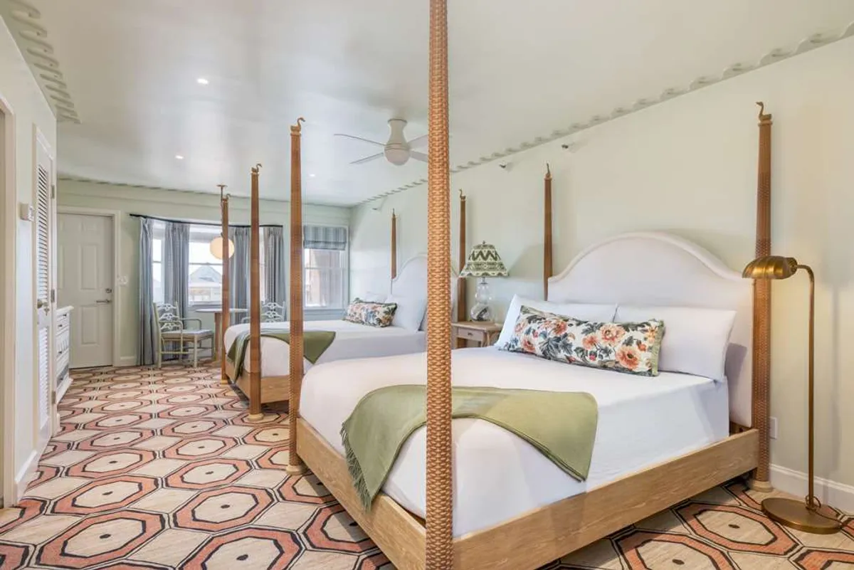 The hero image showcases an inviting studio interior at Sanderling Resort, featuring elegant four-poster beds dressed in luxurious linens, a vibrant patterned rug that adds warmth and charm, a decorative lamp for soft ambient lighting, and a cozy window seat perfect for relaxing or stargazing. Natural light pours in through large windows, creating a bright and comfortable atmosphere ideal for unwinding with family or friends.