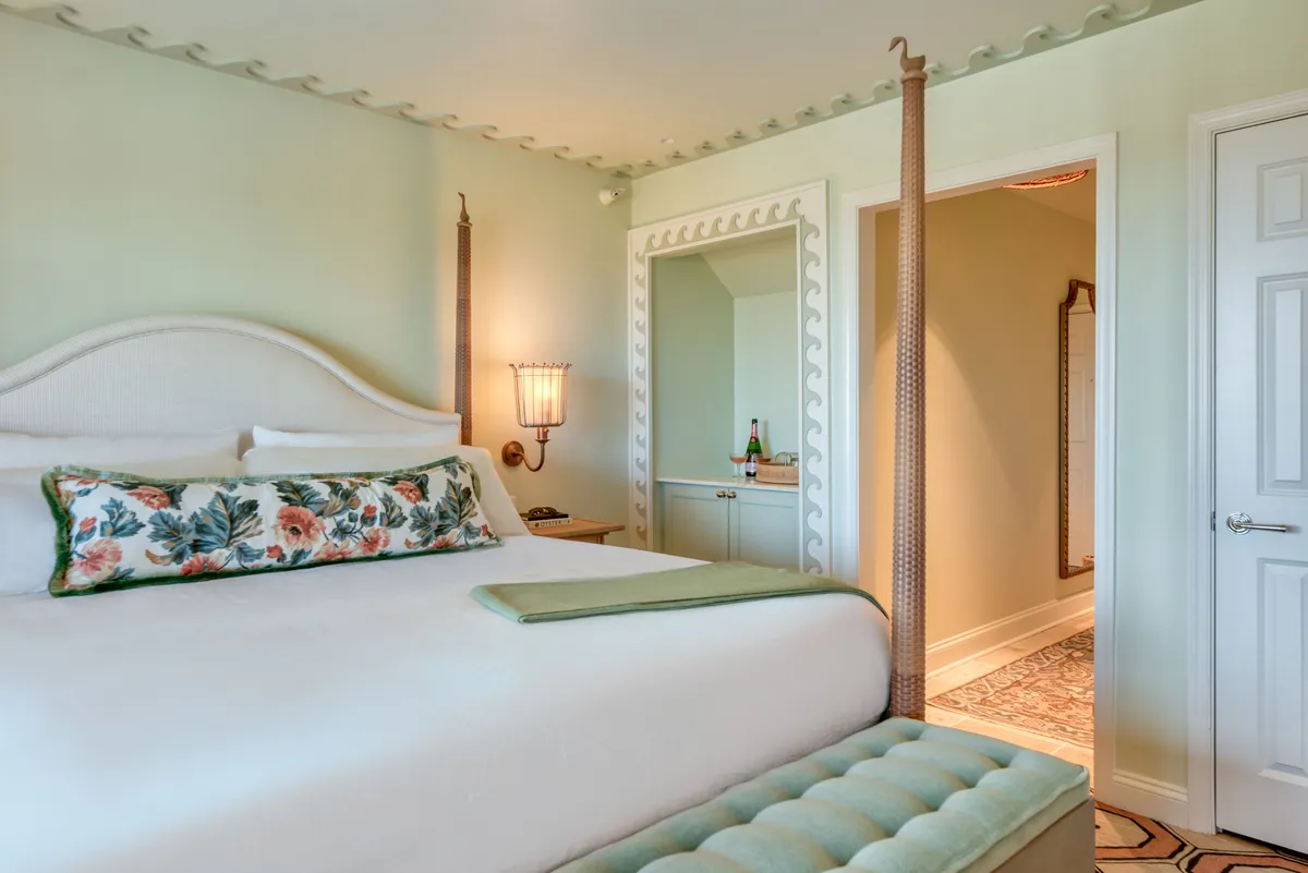 A sunlit studio bedroom featuring a cozy bed adorned with floral pillows, a decorative lamp, and a stylish mirror with an intricate frame. The patterned floor and soft natural light create a peaceful ambiance, making the space inviting for relaxation or reading.