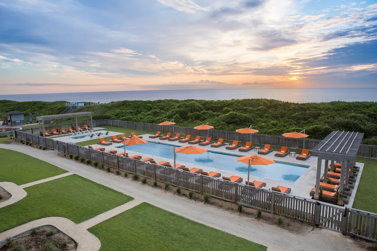 An inviting outdoor pool area featuring sparkling blue water, vibrant orange umbrellas, comfortable lounge chairs, and manicured landscape greenery. The pool sits near the ocean, providing scenic views and a tranquil atmosphere. Groups and families can be seen socializing or relaxing by the water, enjoying both sun and shade.