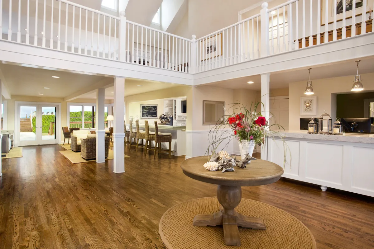 An open-plan living and dining area with a central round table featuring a floral arrangement, connecting seamlessly with large windows that fill the space with natural light. The layout suggests areas for lounging, gathering, and enjoying leisure time within the studio.